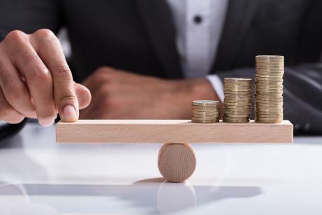 Close-up Of A Hand Balancing Stacked Coins On Wooden Seesaw With Finger Over Desk