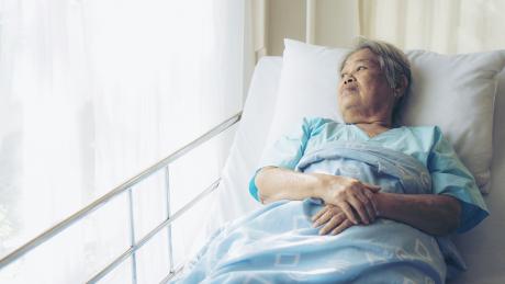older woman alone in the hospital. She is lying in her hospital bed, looking out the window.