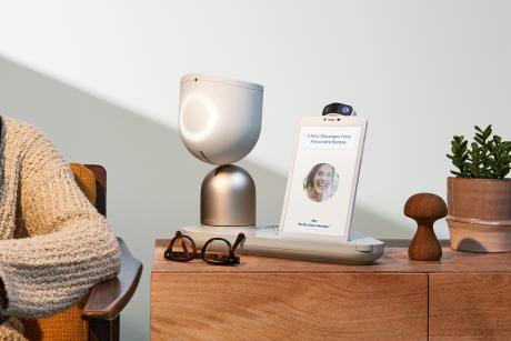 An ElliQ robot sits on a side table. Elbow of older person sitting next to it is seen.