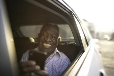 An older Black man in the back seat of a car with the window down smiling, looking at his phone