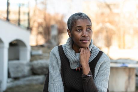 A woman sits outside while looking contemplative. She is dressed warmly in her sweater and looks cozy. She is looking off to the side.