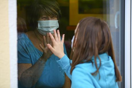Grandmother and granddaughter touch hands on window while on visit. 