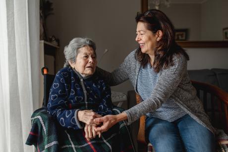 An older Latinx woman sits in a wheelchair next to her daughter. They are holding hands the the daughter is smiling at her.