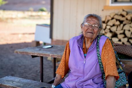 An older indigenous woman sits outside her home in a rural area. She's looking and smiling at the camera.